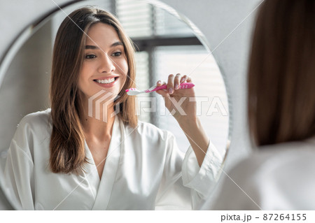 Beautiful Young Woman Brushing Teeth While Standing Near Mirror In Bathroom, Beautiful Young Woman Brushing Teeth While Standing Near Mirror In Bathroom, 87264155