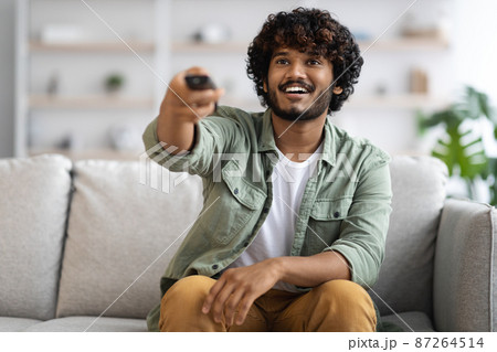 Cheerful indian guy sitting on couch, watching TV Cheerful indian guy sitting on couch, watching TV 87264514