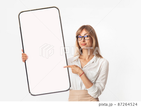 Serious confident young european blond woman in glasses and official wear points finger at big smartphone 87264524
