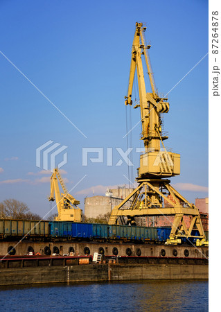 River port crane loading open-top gondola cars on sunny day. Empty river drag boats or barges moored by pier, Empty cars ready for loading. River port crane loading open-top gondola cars on sunny day. Empty river drag boats or barges moored by pier, Empty cars ready for loading. 87264878