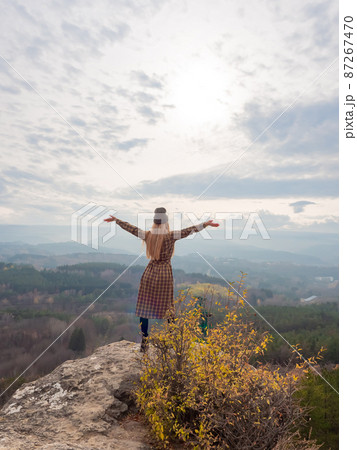 A long haired woman in a dress stands on the edge of a cliff with her arms outstretched and looking into a wooded mountain autumn valley under a cloudy sky 87267470