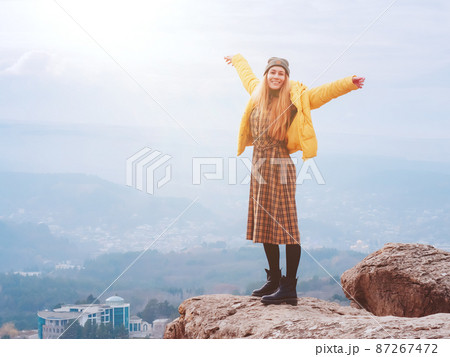 A smiling woman in a plaid dress stands on the edge of a cliff in a smoky hilly valley of the Kislovodsk national park 87267472