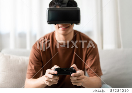 Technology Concept A person wearing a virtual reality headset and holding a black console game while sitting on the sofa 87284010