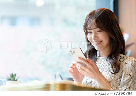 カフェの窓辺の席でスマホを見ながらコーヒーを飲んでいる若い女性 87290794