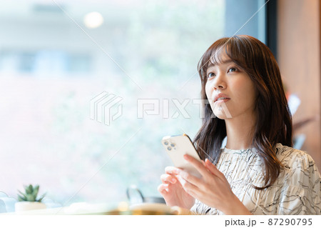 カフェの窓辺の席でスマホを見ながらコーヒーを飲んでいる若い女性 87290795