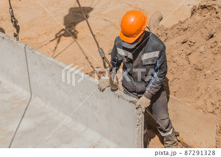 An industrial worker in overalls and a protective helmet attaches a heavy iron hook lifting mechanism with a steel cable rope to a concrete, cement structure An industrial worker in overalls and a protective helmet attaches a heavy iron hook lifting mechanism with a steel cable rope to a concrete, cement structure 87291328