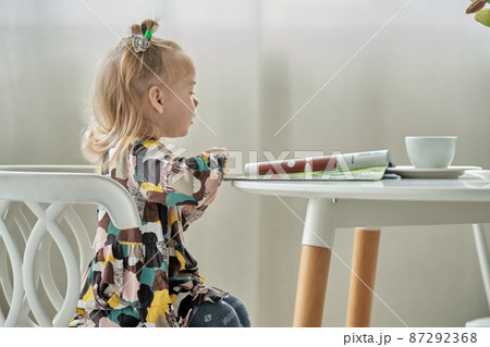 Cute little girl with ponytails on her head reading a magazine sitting on a chair at the table. 87292368