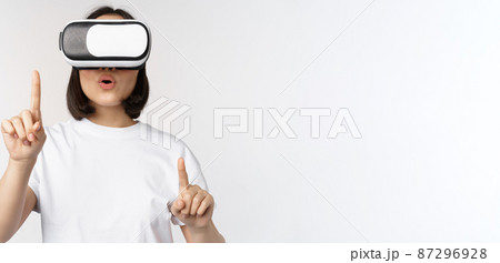 Potrait of asian woman in virtual reality glasses, pointing, choosing smth in VR headset, standing over white background Potrait of asian woman in virtual reality glasses, pointing, choosing smth in VR headset, standing over white background 87296928