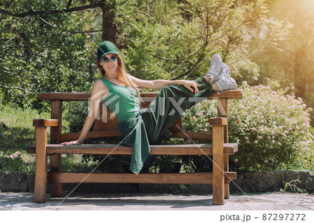 Smiling red-haired girl in a green outfit sits on a bench with her legs raised up in a park under the bright sun Smiling red-haired girl in a green outfit sits on a bench with her legs raised up in a park under the bright sun 87297272