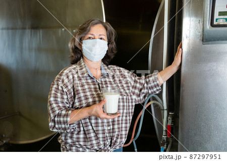 Mature woman in protective mask is holding glass of milk at cow farm Mature woman in protective mask is holding glass of milk at cow farm 87297951