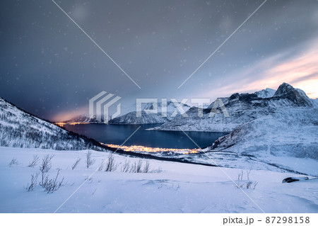 Snow covered mountain range with illuminated town on coastline in blizzard 87298158