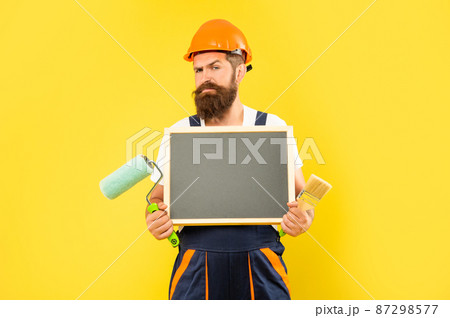 man painter in helmet and work clothes hold paint roller brush and blackboard with copy space 87298577