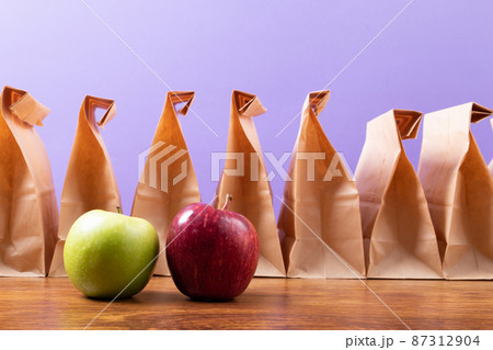 Apple and granny smith apple by row of paper lunch bags on table against purple background 87312904