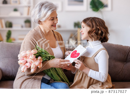 Cheerful little girl giving flowers and handmade postcard to grandmother Cheerful little girl giving flowers and handmade postcard to grandmother 87317027
