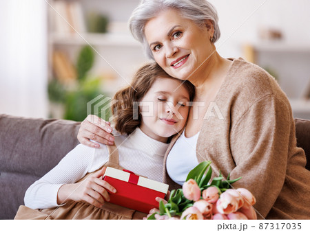 Gently little girl giving flowers and gift to grandmother Gently little girl giving flowers and gift to grandmother 87317035