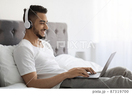 Young Arab Man Wearing Wireless Headphones Using Laptop While Relaxing In Bed Young Arab Man Wearing Wireless Headphones Using Laptop While Relaxing In Bed 87319700