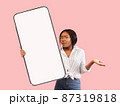 Sad confused young african american female in glasses spreads arms to sides and shows big smartphone 87319818