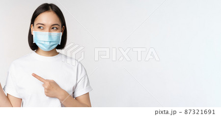 Young korean woman in medical face mask pointing fingers left and looking at logo, showing advertisement or banner, standing over white background Young korean woman in medical face mask pointing fingers left and looking at logo, showing advertisement or banner, standing over white background 87321691