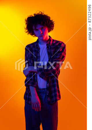 Portrait of young curly man in checkered shirt looking at camera, posing isolated over orange background in blue neon lights Portrait of young curly man in checkered shirt looking at camera, posing isolated over orange background in blue neon lights 87322496