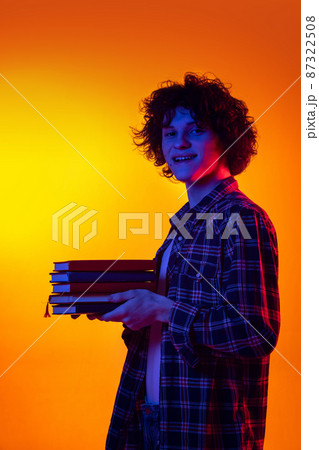 Portrait of young curly man in checkered shirt, student with books posing isolated over orange background in blue neon lights 87322508