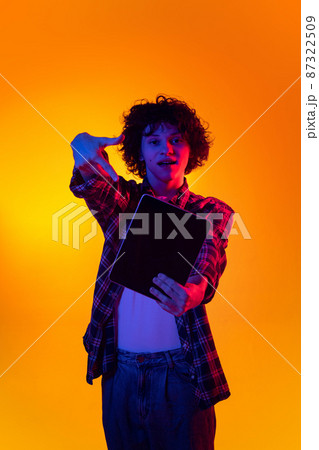 One yougn curly man in checkered shirt pointing at tablet isolated over orange background in blue neon lights 87322509