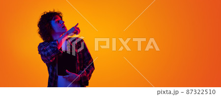 Portrait of young curly man in checkered shirt with tablet pointing right side with interest isolated over orange background in blue neon Portrait of young curly man in checkered shirt with tablet pointing right side with interest isolated over orange background in blue neon 87322510