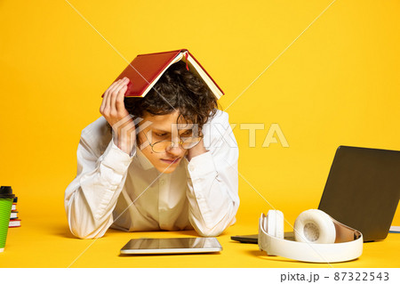 Portrait of young curly man, student in glasses lying on floor around study supplies, studying isolated over yellow studio background Portrait of young curly man, student in glasses lying on floor around study supplies, studying isolated over yellow studio background 87322543