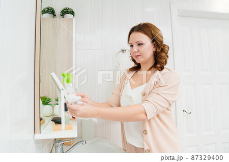 an attractive housewife washes the bathroom mirror with a special glass cleaner. an attractive housewife washes the bathroom mirror with a special glass cleaner. 87329400