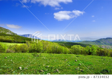 Horizontal background with a magnificent landscape of the Crimean Peninsula. Top view of the mountains covered with lush greenery and the thick clouds below the peaks. Clear blue sky. Copy space. 87334032