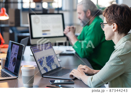 Rear view of businesswoman in eyeglasses sitting at the table and typing on laptop, she analyzing charts online with her colleagues in the background Rear view of businesswoman in eyeglasses sitting at the table and typing on laptop, she analyzing charts online with her colleagues in the background 87344243