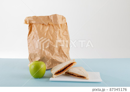 Green apple with peanut butter and jelly sandwich by paper bag at table against gray background 87345176