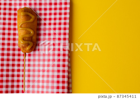 Close-up of corn dog with mustard sauce on checked pattern napkin over yellow background Close-up of corn dog with mustard sauce on checked pattern napkin over yellow background 87345411