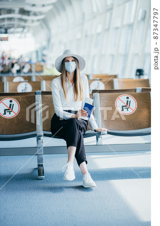 Young woman in a mask sits in an airport lounge during a pandemic and shows her passport and ticket to the camera. 87347797
