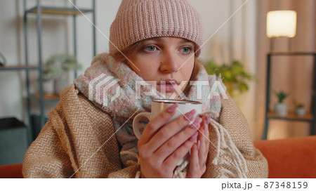 Young sick woman wear hat wrapped in plaid sit alone shivering from cold on sofa drinking hot tea 87348159