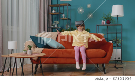Toddler girl sitting on home sofa using virtual reality headset helmet app to play simulation game 87351954