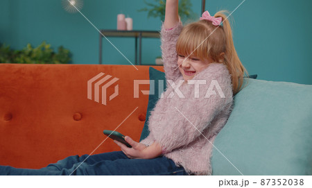 Happy little school kid girl use mobile cell phone doing winner gesture celebrate alone at home sofa Happy little school kid girl use mobile cell phone doing winner gesture celebrate alone at home sofa 87352038