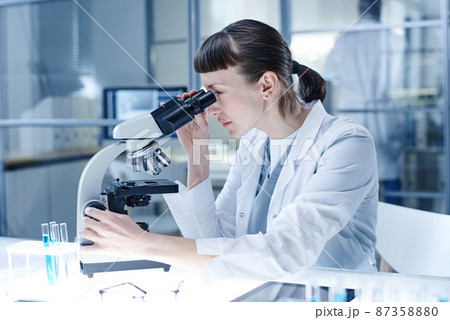 Female scientist in white coat concentrating on her work with analysis, she looking through the microscope at her workplace 87358880