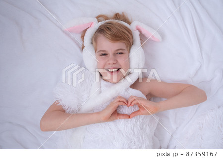 Beautiful little girl plays with rabbit ears. Easter concept. View from above.The child folded the shape of a heart from his fingers. A child on a bed with a fluffy white blanket. Nice and cute Beautiful little girl plays with rabbit ears. Easter concept. View from above.The child folded the shape of a heart from his fingers. A child on a bed with a fluffy white blanket. Nice and cute 87359167