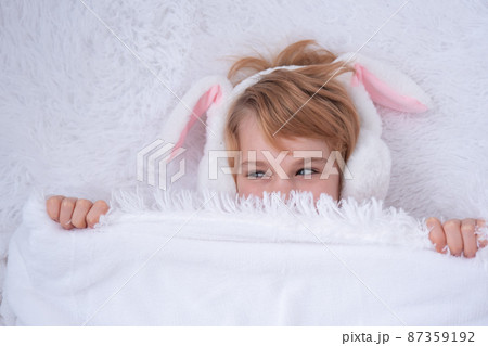 A cute baby appears from under a fluffy white blanket. Teenage girl with rabbit ears. Easter concept. Surprise 87359192
