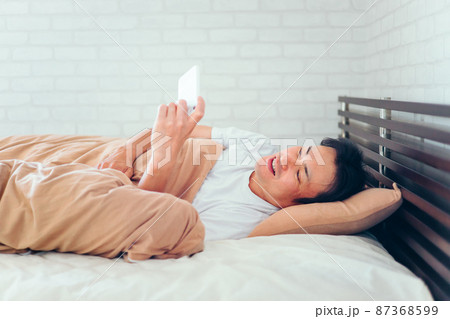 ベッドで寝ながらスマホを見る男性（笑顔） 87368599