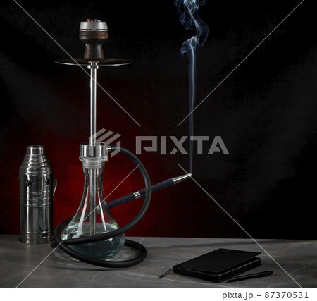 Beautiful modern hookah, from the mouthpiece of which smoke rises, stands on the table, surrounded by a metal protective cap and a diary, on a dark red background. Space for copying. Beautiful modern hookah, from the mouthpiece of which smoke rises, stands on the table, surrounded by a metal protective cap and a diary, on a dark red background. Space for copying. 87370531