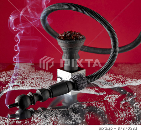 Hookah pipe, curved in a spiral, from the mouthpiece of which smoke flows against the background of a ceramic hookah bowl with a small stand on a scarlet background with reflections. Hookah pipe, curved in a spiral, from the mouthpiece of which smoke flows against the background of a ceramic hookah bowl with a small stand on a scarlet background with reflections. 87370533