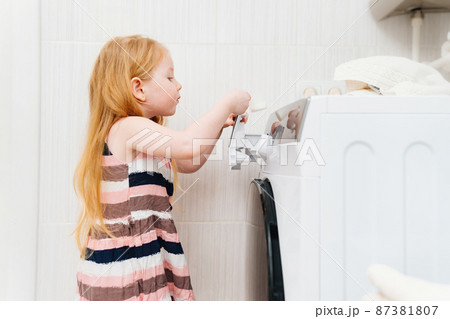 a cute little girl puts washing powder in the washing machine.  87381807