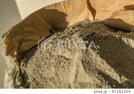 Grey sand-concrete dry mix in an open paper bag. Concrete dry powder for leveling walls and floors. Repair in the house with your own hands. DIY. Selective focus Grey sand-concrete dry mix in an open paper bag. Concrete dry powder for leveling walls and floors. Repair in the house with your own hands. DIY. Selective focus 87382304
