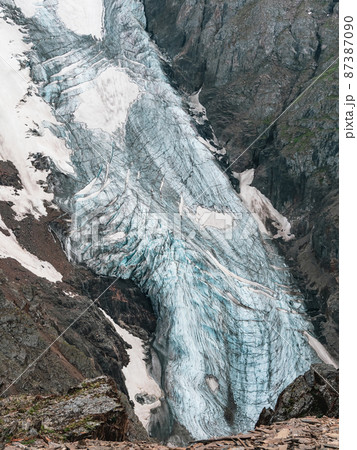 Vertical mountain landscape with blue long vertical glacier tongue with cracks among rocks. Aerial view to large glacier with icefall. Nature texture of mountain glacier with fissures close up. 87387090