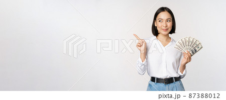 Smiling young modern asian woman, pointing at banner advertisement, holding cash money dollars, standing over white background Smiling young modern asian woman, pointing at banner advertisement, holding cash money dollars, standing over white background 87388012
