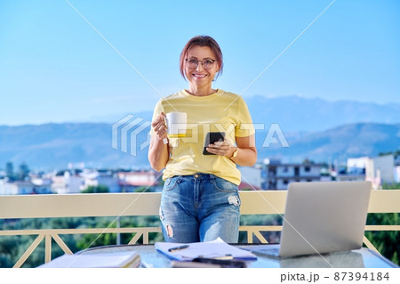Middle-aged woman working at home on terrace, relaxing with mug of tea and smartphone 87394184