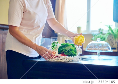 Fresh vegetables in mesh eco bag on table in kitchen, woman puts vegetables on plate Fresh vegetables in mesh eco bag on table in kitchen, woman puts vegetables on plate 87394185