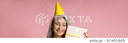 Happy mature Asian lady with yellow party hat holds gift box on pink background 87401869