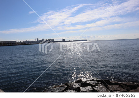 海岸の岩とキラキラ光る水面と青い海面の海と港の突堤と人工島の街並みと雲と青空の見える風景 87405565